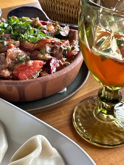 A table in Marneuli, Georgia, holds a feast: a clay pot filled with a stew of tomatoes, meat (possibly lamb or beef), and herbs; a plate of dumplings; and an amber-colored wine glass. A woven basket is slightly visible at the far left edge. The scene is indoors, likely a restaurant.