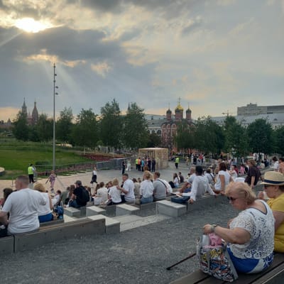In a scene set in an outdoor amphitheater with tiered seating, a diverse group of people are gathered, some seated and others standing. The architecture suggests a park or public space, with a grassy area and trees visible in the background. A prominent religious building with golden domes and red-brick walls is situated in the middle ground, hinting at a historical or cultural landmark. The sky above is partly cloudy, with rays of sunlight breaking through, creating a dramatic effect. A light pole with multiple lights stands tall, adding to the urban landscape. Several individuals are interacting, some engaged in conversation while others observe the surroundings. The presence of a security guard in a yellow vest indicates a managed event or public gathering. The overall atmosphere appears relaxed and social, with people enjoying a pleasant day outdoors. The city of Путрышкаўскі сельскі Савет, Belarus, is not visually represented in the image, nor can it be inferred from the visual cues present.