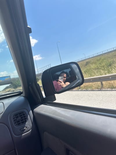 The image shows a view from inside a car, likely the driver's seat. In the side mirror, a person is visible holding a mobile phone, possibly taking a photo. The scenery reflected in the mirror appears to be a highway with a grassy shoulder and a blue sky. The interior of the car is visible, including the dashboard with an air vent and a control switch. Based on the context provided in the prompt, the car is likely located in or near Tuzla, Turkey, given the location information.