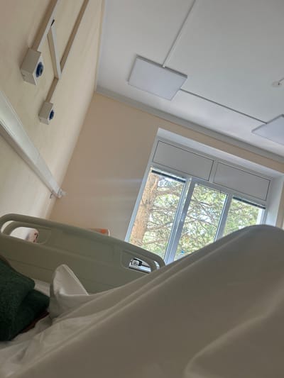 The image depicts the interior of a hospital room, with a focus on a hospital bed and a window. The bed is made with white linens, and a dark green pillow is visible on the left side. The hospital bed rails are a light beige color. To the right, a large window with multiple panes offers a view of trees and foliage, suggesting an outdoor environment.
On the wall to the left of the bed, there are two white electrical outlets with blue connectors, mounted on a beige wall. Above the window, the ceiling features recessed square light panels. A white cable is visible extending from the wall near the bed towards the window.
The room appears clean and functional, typical of a medical facility. The lighting is soft, likely from the overhead panels and natural light from the window. There are no people directly visible in the foreground, though a leg with an orange band is partially visible under the sheets on the bed. The overall impression is one of a patient's room within a healthcare setting.
The location provided, Ливадийский поселковый совет, Ukraine, suggests this is a hospital in that administrative region. However, the visual elements themselves do not provide specific geographical markers beyond the general appearance of a hospital room and the view through the window.