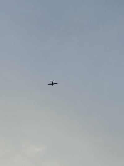 This media file shows a small airplane in silhouette against a pale, overcast sky. The airplane has a single propeller and appears to be a light aircraft. It is positioned in the upper-middle portion of the frame. The sky is a uniform, muted blue-grey, suggesting it might be late afternoon or early morning, or possibly a day with significant cloud cover. There are no discernible landmarks or location cues within the frame, beyond the implied open sky. The image is still and there is no indication of any activity or event beyond the plane's presence in the sky. There is no visible text in the image. The context of Nowy Dwór Mazowiecki, Poland, suggests the plane is likely flying over or near this area.