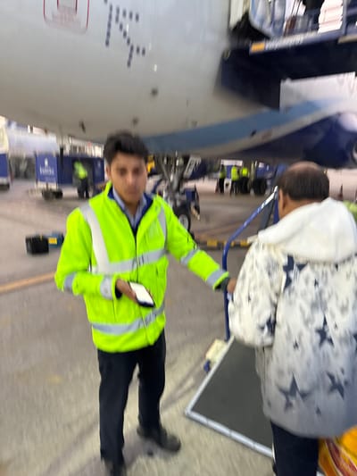 A man in a neon yellow jacket and black pants stands next to an airplane, holding a white smartphone. He is facing the camera with a slight smile. Behind him, two other individuals are boarding the plane using a mobile stairway. Luggage carts and other ground support equipment are also visible near the aircraft. The overall setting appears to be an airport tarmac during daylight.