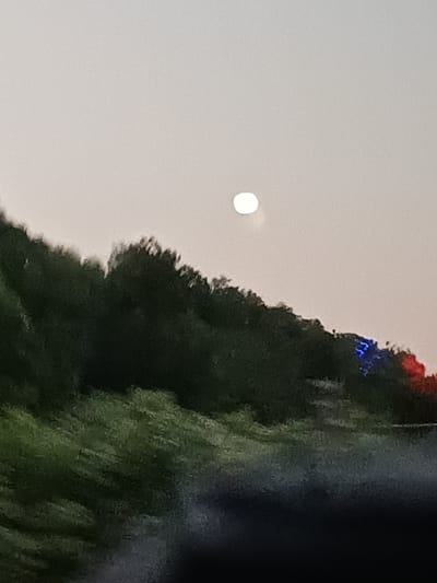A bright white orb, possibly the moon, is visible in the hazy, pale sky above a dark, undulating line of trees. The trees appear dense and dark green, with some reddish and bluish lights showing faintly on the right side of the treeline. In the foreground, a blurry, green, and dark area suggests a moving perspective, perhaps from a vehicle. The scene is likely an outdoor setting in Adlerskiy, Russia, given the context of the city.