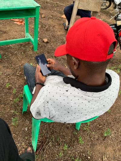 A high-angle, outdoor medium shot shows a dark-skinned man wearing a red baseball cap and a light gray shirt with a black speckled pattern sitting on a green plastic chair. He is holding a dark-colored smartphone in his right hand and appears to be looking at the screen. He is also wearing a black beaded bracelet on his left wrist. In the background, a green metal table with a shelf is visible, along with dry leaves and twigs on the ground. To the right of the man, part of a motorcycle is visible, suggesting an urban or semi-urban setting, possibly in Tabu, Nigeria, given the context provided. Another person wearing dark pants and sandals is seated behind a wooden structure, further to the left. The ground is composed of dirt and scattered debris.