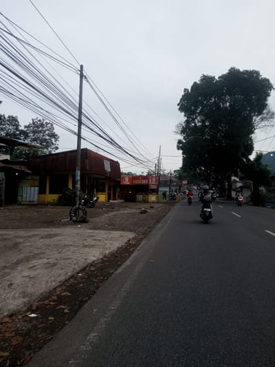 The image displays an outdoor street scene in Sat Thay (East), Myanmar, under an overcast sky. An asphalt road extends from the foreground into the distance on the right. To the left of the road, a concrete and dirt shoulder is present. A tall utility pole, laden with numerous black overhead wires, stands in the left foreground. Behind the pole, a series of single-story commercial buildings are visible. The closest is a yellow structure with a rusty corrugated metal roof, adjacent to an orange-fronted establishment displaying a "BAKSO SOLO" sign. Several motorcycles are parked near these buildings. On the road, multiple individuals are riding motorcycles away from the viewer; some are observed wearing helmets. Large, dark-foliage trees line the right side of the road further in the distance. Two discarded vehicle tires are located on the ground near the base of the utility pole.