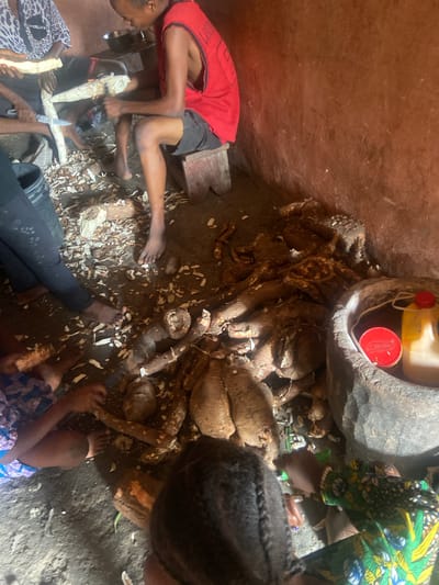 In the city of Nagazi, Nigeria, a group of individuals is engaged in preparing what appears to be cassava. The scene is set in a rustic, earthen-walled interior, possibly a dwelling or food preparation area. Several large, fibrous cassava roots are visible on the ground, some partially peeled and others still intact. Peels and trimmings litter the dirt floor, indicating an ongoing process.

One person, seated on a wooden stool, is actively peeling a cassava root with a knife. This individual wears a red sleeveless shirt and dark shorts. Another person, partially visible in the upper left, is also peeling cassava, holding a knife and a large piece of the root. A third individual, in the lower left, is wearing dark trousers and appears to be interacting with the cassava or assisting in the preparation. In the foreground, the back of a person with braided hair is visible, and to the right, another person in a colorful patterned garment is leaning over, also seemingly involved in the task.

A large, rough-hewn wooden mortar sits in the right side of the frame, containing two containers: a yellow plastic bottle with a red cap and a red cylindrical container. The overall environment is dimly lit, with the earthy tones of the walls and floor dominating the scene. The focus of the activity is the preparation of the cassava, suggesting a communal effort for food processing.