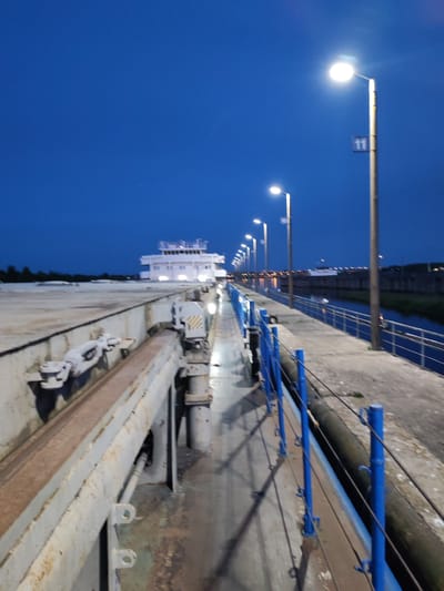 The image shows a view of a canal lock in Sheksna, Russia, at dusk or night. A large, white, multi-story vessel is docked inside the lock, illuminated by interior lights. The lock walls are made of concrete, with industrial-looking metal fixtures and railings. Lampposts line the walkway along the lock, casting long shadows on the ground. The sky is a deep blue, indicating twilight or a clear night sky. The overall impression is one of industrial infrastructure and waterway transport.