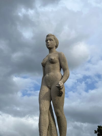 A full-body, low-angle, medium shot captures a stone statue of a nude woman against a cloudy sky. The statue stands in a park or public space in Deauville, France. The statue's rough texture and carved features are visible, with its head tilted slightly to the left and its body facing forward. The woman's left arm is bent at the elbow, with her hand resting near her hip, holding a small object. Her right arm hangs down by her side. The sky is filled with a mix of dark and light gray clouds, with patches of blue visible in between. The overall impression is one of a weathered, classical sculpture set against a dramatic, natural backdrop.