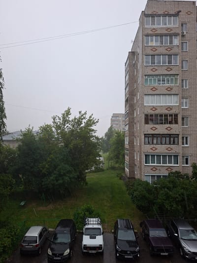 In Murom, Russia, a residential courtyard is depicted during a rain shower. The scene features a multi-story brick apartment building on the right, with multiple windows and balconies. In the foreground, several parked cars are visible on a paved surface, including a silver hatchback, a black minivan, a white SUV, a dark purple SUV, and a silver sedan. Beyond the cars, a grassy area is interspersed with trees and bushes. In the background, another apartment building and some foliage are present. The sky is overcast, contributing to a dull, grey ambiance.