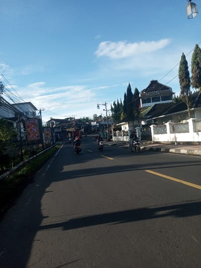 The image displays a street scene in Khouathineung, Laos, viewed from a low angle, under a clear blue sky with sparse white clouds. The asphalt road, marked with a solid yellow line on its right, curves gently to the left in the distance. Three motorcycles are visible on the road, traveling away from the viewer; the closest on the right is a dark-colored motorcycle with a single rider wearing dark clothing and a helmet, followed by another dark motorcycle, and a red motorcycle further back on the left. Long shadows, indicative of a low sun angle, are cast across the road. The left side of the road features a narrow, unpaved shoulder, a low concrete wall, dense green foliage, utility poles with numerous overhead wires, and various buildings, including one with a large banner depicting a person's face. The right side of the road presents a concrete sidewalk, a white-painted fence, a row of tall, slender coniferous trees, and a multi-story building with a distinctive dark, multi-tiered roof. A street light fixture hangs partially visible in the upper right corner.