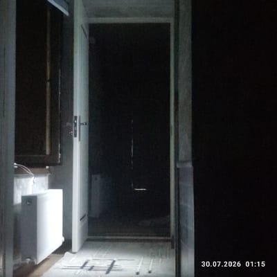 The image shows an interior hallway with a white door ajar, revealing a dark room beyond. To the left, a white radiator is visible, along with some wires and a window frame. The floor is tiled with a light-colored pattern. The date and time stamp "30.07.2026 01:15" is present in the lower right corner. The scene appears to be a domestic setting, possibly an apartment or house in Uchqoʻrgʻon, Uzbekistan. There are no people or animals visible in the image.