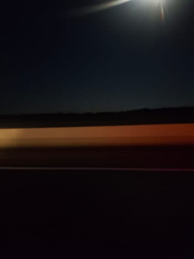 The image depicts a blurred, horizontal streak of orange and yellow light against a dark blue and black background. This light streak appears to be a long exposure photograph of moving lights, likely from vehicles on a road. Above the light streak, a faint horizon line of dark hills or mountains is visible. At the top of the image, a bright, sun-like glare with rays suggests a light source, possibly the moon or a distant artificial light, casting a beam downwards. The lower portion of the image is dark, with a thin white line visible, potentially a road marking. There are no discernible people, specific objects, or actions visible due to the blur and darkness. The scene implies a nighttime setting, possibly on a highway or open road. The location in the city of Mingchinar, Uzbekistan, cannot be determined from the visual information present in the image.
