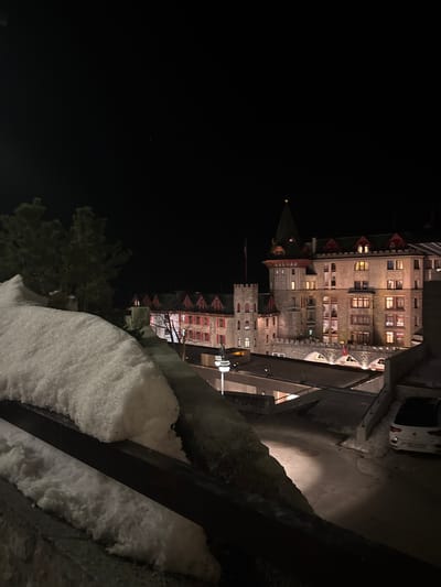 The image depicts a nighttime winter scene in Sankt Moritz, Switzerland, specifically featuring the Badrutt's Palace Hotel. In the immediate foreground, a dark, snow-covered parapet or railing extends across the lower left quadrant, with a significant accumulation of granular white snow topping a dark horizontal bar. Behind this, to the left, several dark evergreen trees are visible against the dark sky.

Below the foreground railing and extending into the mid-ground, a multi-level concrete and stone architectural complex descends. A winding, partially snow-covered asphalt road or pathway with visible tire tracks is illuminated by artificial light, revealing patches of asphalt. A white, modern hatchback vehicle is parked on the right side of this pathway, facing away from the viewer, with its rear visible.

The dominant structure in the background is the Badrutt's Palace Hotel, a large, multi-story building constructed of stone with ornate architectural details. It features a prominent green conical-roofed tower with a finial, several smaller turrets, and numerous windows, many of which are illuminated with warm yellow light. Red-shuttered windows are visible throughout the structure. A flag pole with a dark flag is affixed to one of the smaller towers on the left side of the hotel. Below the main hotel facade, a series of stone arches form part of its base or an integrated bridge structure, with a red flag featuring a white cross visible beneath one arch. The sky above is uniformly dark, indicating a clear night. No individuals are visible.