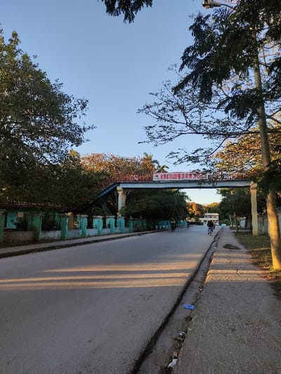 An upward-angled perspective displays a paved road flanked by trees and structures. A prominent sign spans above, adorned with red Chinese lettering. Sunlight casts long shadows across the pavement. Lush green foliage lines one side, while a pathway with a darker curb runs along the opposite edge. In the distance, a white vehicle is visible. The sky is clear and blue.