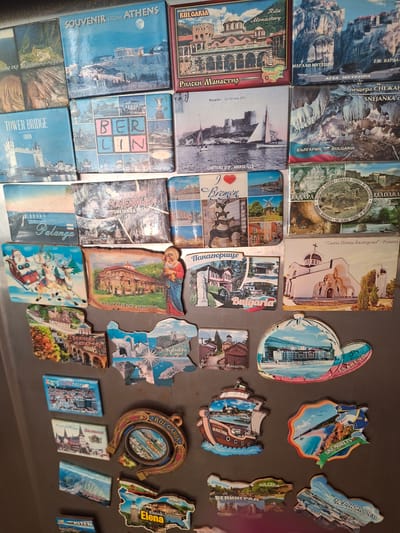 The image displays a collection of refrigerator magnets, primarily featuring landscapes, architecture, and some illustrations. The magnets are arranged on a dark, metallic surface, likely a refrigerator door.
The subjects include diverse scenes such as natural landscapes with mountains and water bodies, historical buildings like monasteries and churches, and cityscapes. Some magnets feature illustrative elements, like one with a sleigh and reindeer, suggesting a festive or winter theme. Another magnet depicts a horseshoe with "BULGARIA" and "KRANEVO" visible. There are also magnets with maps of coastal regions and notable landmarks.
No specific activity or event is taking place as the image is a static display of objects.
Notable details include the presence of text on many magnets, providing place names, often in Cyrillic script, such as "RILA MONASTERY," "BREMEN," "BERLIN," "TOWER BRIDGE," "ATHENS," "TSAREVO," "SINEMORETS," "BALCHIK," "VENECIA," "KRANEVO," and various Bulgarian locations like "KALAKARA CAPE," "ELENA," and "KARLOVO." The magnets vary in shape and size, with some being rectangular, others irregular outlines of the depicted locations, and one in the shape of a horseshoe. The colors are generally vibrant, indicating printed images on the magnet surface. The overall impression is a personal collection of travel souvenirs. The backdrop appears to be a kitchen or similar domestic setting, given the context of refrigerator magnets.