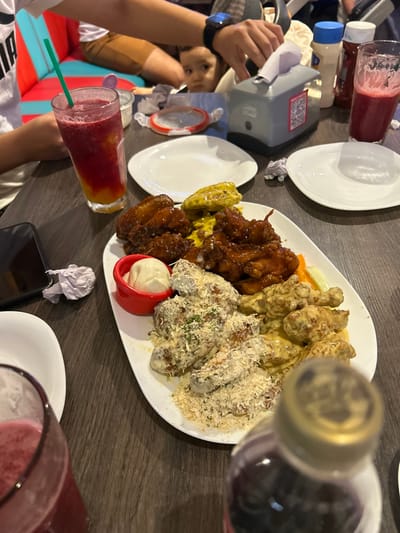 A table displays a platter of chicken wings prepared in various styles, ranging from saucy to parmesan-covered, accompanied by carrot and celery sticks, and a small bowl of white sauce. Drinks, including a glass of layered red and yellow liquid with a green straw, and plates are placed around the platter. A cell phone, napkins, and other containers are also visible. A child is seated at the table with an adult.