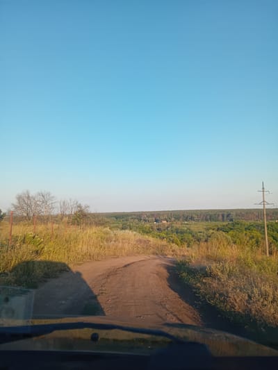 The image shows a view from a vehicle's windshield looking out at a dirt road that curves to the left and continues into a landscape of rolling hills covered in trees and vegetation. The sky is clear and blue. In the foreground, the shadow of the vehicle is cast on the road and to the left, obscuring the ground. To the left of the road, there is dry grass and some bare trees, possibly a fence line made of posts. In the distance, a power line pole is visible on the right side of the frame, and the horizon is lined with a dense forest. The scene is likely in a rural area given the dirt road and natural landscape. There are no people or animals visible in the image. The lighting suggests it is daytime, likely late afternoon due to the long shadows. The location is identified as Kochetok, Ukraine.