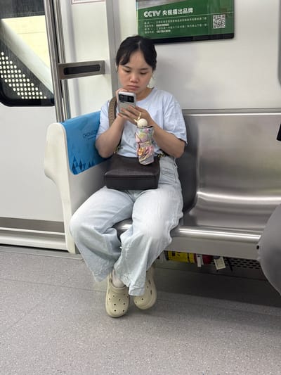 A young East Asian woman sits on a silver train seat, holding a smartphone. She is wearing a light blue t-shirt, light wash baggy jeans, and beige Crocs with star-shaped cutouts. A dark brown leather purse hangs from her shoulder, and she has a small, colorful stuffed toy in her hand, resting on her phone. The train car features a blue and white seat back with a repeating diamond pattern and the word "CHONGQIN" visible. A metal handrail is mounted on the wall, and a window with illuminated dots is to the left. A fire extinguisher is visible under the seat. The flooring is speckled grey. The image is taken indoors, likely in a public transportation setting.
