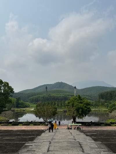 The photograph, taken in Thủy Xuân Ward, Vietnam, presents a landscape view. In the foreground, stone steps and pavement stretch towards the right side of the frame. A body of water, likely a lake or river, is bordered by lush green trees and vegetation. On the opposite bank of the water, a series of hills and mountains recede into the distance, partly obscured by a light haze. A tall, slender structure resembling a monument or tower stands atop one of the hills. Several individuals are walking along the paved area beside the water, some dressed in bright yellow clothing. The sky above is partly cloudy with patches of blue visible.