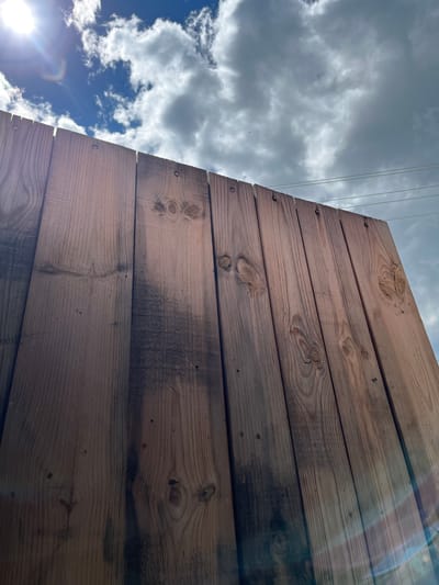 A low-angle outdoor shot of a wooden fence with a cloudy sky and a bright sun in the background. The fence is made of vertical wooden planks with visible grain and knots. There are some dark stains on the planks. The sky is blue with scattered white and gray clouds. Power lines are visible in the upper right corner. The sun is shining brightly from the upper left, creating lens flare. The image was taken in Abzakovo, Russia.