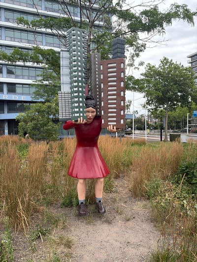 This media file shows a statue of a woman lying down, holding onto a segment of a building facade. The statue is positioned outdoors, with a gravel path to its right and tall, dry grass on its left. Behind the statue, a modern building with many windows is visible, along with trees and a street with a few cars. The sky appears overcast, suggesting a cloudy day. The statue is wearing a red dress and brown shoes with grey socks. The woman's face is visible, with a slight smile. The building segment she is holding is brown with a pattern of white rectangles. A second, green building segment with a grid pattern is also attached to the statue. In the background, the letters "RDE" are visible on the side of the modern building, and smaller text that reads "Bogaz d'len 15-57" can be seen near the base of the building. The location is Rijswijk, Netherlands.