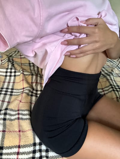 A person is lying on a plaid blanket, wearing a pink hoodie and black shorts. Their hand is holding the edge of the hoodie, revealing their midriff. The scene is captured from a close-up, overhead perspective, focusing on the torso and immediate surroundings. There is no identifiable information about the location being Basarabeasca, Moldova.