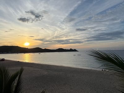 This is a wide-angle, eye-level shot of a tranquil beach at sunset in Song Cau, Vietnam. The sun, a bright orb, is low on the horizon, casting a warm golden glow across the sky and water. The sky is a canvas of wispy clouds in shades of white, gray, and blue, with streaks of orange and yellow near the sun. Rolling hills form a dark silhouette against the vibrant sky, punctuated by a few scattered lights indicating a small settlement.
The beach itself is wide and sandy, with gentle waves lapping at the shore. The water is calm and reflects the colors of the sunset. Small boats are dotted across the calm sea, some near the shore and others further out. A few people are visible on the beach; two figures sit on the sand near the water's edge, and another person stands by the water. In the foreground, the dark fronds of a palm tree create a natural frame for the scene. A red vehicle with a camper shell is parked on the left side of the beach, and what appears to be a small, solitary boat is pulled up on the sand further down the shoreline. The overall atmosphere is peaceful and serene, capturing the beauty of a coastal sunset.