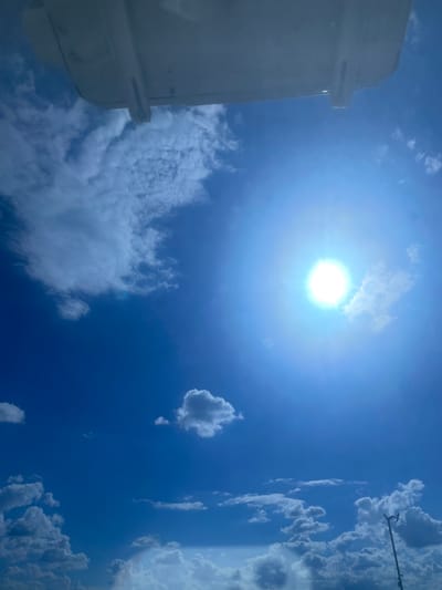 The image depicts a bright, sunny sky with scattered white clouds. The sun is prominently visible in the upper right quadrant, emitting a strong glare. A portion of a white, rectangular structure, possibly the underside of an aircraft or a building overhang, occupies the top portion of the frame. The lower part of the image shows a landscape of clouds, with a faint hint of a rainbow visible in the bottom left corner. A tall, thin object with two lights on top, resembling a street lamp or a fixture, stands on the right side of the frame. The scene appears to be an outdoor environment. There is no information in the image to indicate the location is Podkletnoe, Russia.