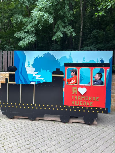 In this photo, two individuals are posing in a cutout of a train carriage, part of an outdoor attraction. A man in a dark blue jacket looks down, while a boy in an orange shirt and a baseball cap peers out from a lower window. The backdrop is a vibrant blue scene depicting stylized trees and mountains, with a white heart symbol and Russian text reading "Я ГУАМСКОЕ УЩЕЛЬЕ" (I love Guamka Gorge) visible on the red part of the train. The train itself is painted black with decorative yellow circles. The scene appears to be set in a park or recreational area, with cobblestone pavement visible to the right. The lighting suggests it is daytime.