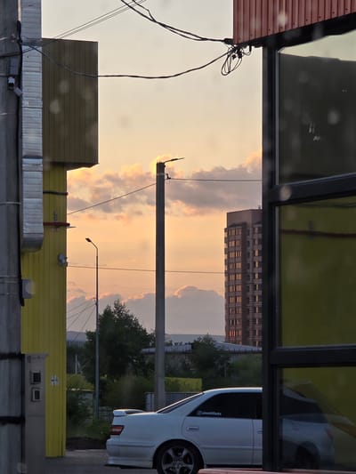 The image, captured in Chita, Russia, shows an urban scene at sunset. The sky is filled with warm hues of orange and yellow, with scattered clouds illuminated by the setting sun. In the foreground, there are elements of urban infrastructure, including what appears to be a corrugated metal roof on the left and a yellow structure on the right, both reflecting the sky's colors. Wires and a utility pole are also visible, suggesting the presence of electrical infrastructure.
In the middle ground, a tall, multi-story building with many lit windows stands against the sunset, hinting at a residential or office building. To the right, a white car is parked, its reflection visible in a mirrored surface above, possibly a window or a display. The car has a sticker on its rear window that reads "@aimettilazta".
The overall impression is one of a typical evening in a city, with the natural beauty of the sunset contrasting with the man-made structures. The lighting suggests it is late afternoon or early evening. There are no people clearly visible in the image. The weather appears to be clear apart from the clouds.