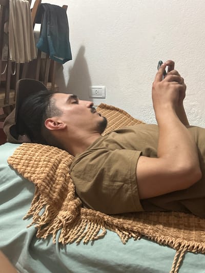 An adult male is observed lying on his back on a bed covered with a light blue sheet, his head turned to the right. He has dark hair, styled with a fade and pulled back, a mustache, and a short beard. He wears a plain, short-sleeved, olive-drab t-shirt. A textured, light brown woven blanket with fringes partially covers him and the bed. He holds a dark-colored smartphone with both hands, appearing to interact with it, his eyes directed towards the device. Behind his head, a dark hat rests on the bed near the wall. The background consists of a plain white wall, on which a white Type I electrical outlet is mounted. To the upper left, wooden furniture, possibly a chair, is visible with light and dark teal garments draped over it. Shadows are cast on the wall by the furniture and the individual's head, suggesting an indoor setting with artificial lighting. The power outlet type is consistent with those used in Mar del Plata, Argentina.