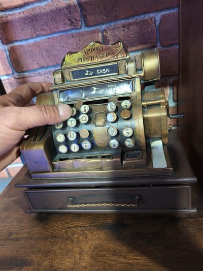 An overhead view depicts a human hand interacting with an antique-style cash register situated on a dark wooden surface in Picassent, Spain. The cash register features a bronze and dark brown metallic finish with visible wear. An elevated sign at the top reads "AMOUNT PURCHASED" and "CASH". Below this, a mechanical readout displays "Dollars" and "Cents" with visible numbers. The device incorporates multiple circular keys; some are numeric (e.g., 5, 9, 7, 3, 8, 4), others alphabetic (P, T, Q, U, I, E), and some bear symbols (+, &). A light-skinned human thumb is observed pressing a key marked 'P' on the left side of the machine. The cash register rests on a dark brown wooden surface exhibiting distinct grain patterns. A single drawer with a light brown twisted metallic handle is positioned beneath the main unit. The background consists of a wall constructed from reddish-brown bricks with light-colored mortar. A small, rectangular white adhesive strip is visible on the lower right side of the cash register, adjacent to a black crank handle extending from the machine's right side.