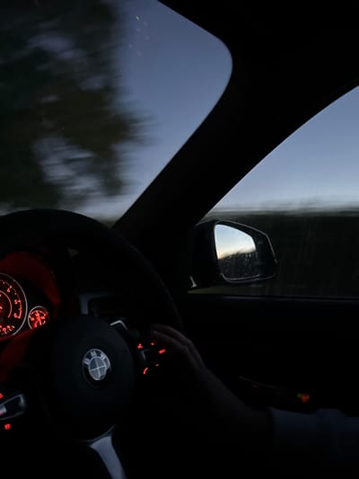 The image shows the interior of a car at dusk or dawn, as evidenced by the dim lighting and the sky visible through the windshield and side window. The dashboard gauges are illuminated with red light, indicating some level of activity within the car. A hand is visible on the steering wheel, which features a BMW logo, suggesting the vehicle's make. The driver appears to be operating the vehicle, with the side mirror reflecting the twilight sky and a dark landscape. The scene suggests movement, likely on a road in a rural or semi-rural area given the dark, indistinct surroundings. The location is stated to be North Hertfordshire, United Kingdom, but no specific landmarks are identifiable within the image to confirm this.