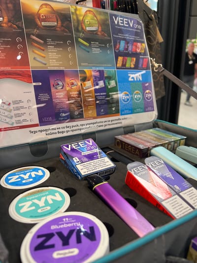 This image captures a retail display of various nicotine and vaping products in an indoor commercial setting, likely a store or kiosk in Parvomai, Bulgaria. The display consists of a black tray or case, holding different brands and types of items, alongside an informational sheet.
Prominently featured on the right side of the tray are several boxes of "VEEV one" and "VEEV PRIME" vaping devices or pods, including "Purple Wave" and "Red" varieties. A purple "VEEV" disposable vape pen is also visible. Below these, three round white tins of "ZYN" nicotine pouches are displayed, with labels indicating "Cool Mint Mini," "Spearmint Mini," and "Blueberry" flavors, along with nicotine strengths. In the middle-top, stacks of "OTEREA" heated tobacco sticks are arranged.
The left side of the image is dominated by an open, laminated information sheet. This sheet presents details on product lines such as "VEEV one," "TEREA," "LEVIA," and "ZYN," listing various flavors like "Teak," "Amber," "Yellow," "Silver," "Soft Fuse," "Turquoise," "Summer Pearl," "Berry Pearl," "Exotic," "Dark Berry," and different "Classic" tobacco options. The visible text on the packaging and the information sheet is primarily in Bulgarian, including health warnings such as "Това изделие съдържа никотин, който води до силно пристрастяване. Не се препоръчва за употреба от непушачи." (This product contains nicotine, which is highly addictive. Not recommended for use by non-smokers.) and "Само за пълнолетни." (For adults only). A blurred reflection of a person is visible in the background, and a handbag hangs in the upper left corner.