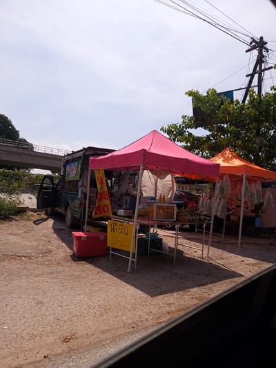The image shows a roadside scene in Hua Hin City Municipality, Thailand. A dark green pickup truck, modified to serve as a mobile vendor stall, is parked on a dirt patch next to a paved road. The driver-side door is open. Two pop-up tents, one pink and one orange, are erected next to the truck, indicating a marketplace setup. The tents appear to house food items. A red cooler sits on the ground near the truck. A yellow sign with Thai script and the numbers "10-20" is visible near the pink tent.
In the background, a concrete overpass is visible, and a utility pole stands with multiple wires. The sky is bright with scattered clouds, suggesting daylight conditions. There is no evidence of pedestrian or vehicular traffic nearby.