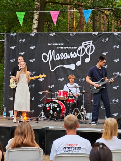 An outdoor music performance is taking place on a stage at night. A band of four musicians is on stage. A female singer in a white dress is at the front, singing into a microphone. To her left, a male musician is playing a bass guitar. To the right of the singer, a male guitarist is playing an electric guitar. In the background, a male drummer is playing a drum set. The stage is adorned with a black banner with a white logo. Triangular flags in green, pink, and blue are strung across the top of the frame. The audience is seated in chairs in front of the stage, facing away from the camera. The setting appears to be a park or outdoor event space. The city of Ракітніцкі сельскі Савет, Belarus is not visible in the image.