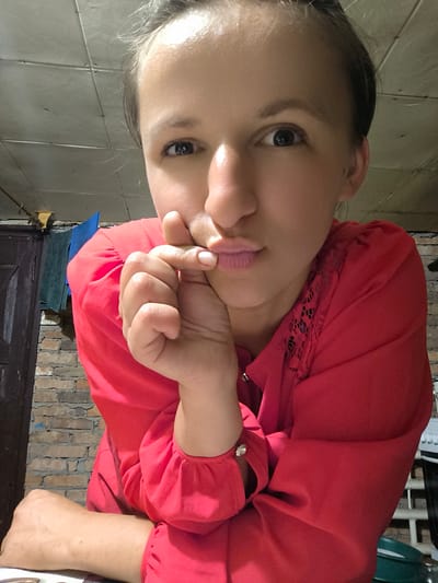 This is a close-up, point-of-view photo of a woman from Anastasivka, Ukraine, who is leaning forward and resting her chin on her hand. She has fair skin, dark eyes, and light brown hair pulled back. Her lips are slightly pursed, and she is looking directly at the camera with a neutral expression. She is wearing a bright red blouse with lace detailing on the shoulders and button cuffs.
The background is a brick wall and a textured ceiling, suggesting an indoor setting. To the left, a dark wooden door and some hanging towels are visible. The lighting appears to be artificial, possibly from an overhead light, and it casts shadows that highlight the contours of her face. The overall impression is casual and intimate, as if she is taking a selfie in her home. No text is visible in the image.