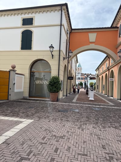 This image depicts a picturesque outdoor pedestrian shopping street in Noventa di Piave, Italy, characterized by a series of upscale boutiques. The view looks down a walkway paved with grey and brown bricks laid in a herringbone pattern, bordered by white lines.
On the left, a light-colored building features an arched storefront with "ARMANI" clearly visible in white text on a lowered dark shutter. A potted green bush sits in front of the window. Further along this side, another building displays arched window frames and dark shutters. On the right, a building with a warm terracotta-orange facade also features multiple arched openings, with "FENDI" visible on a sign above one. In the distance, a white standalone sign with "GIVENCHY" is partially visible.
The architecture suggests a modern interpretation of traditional Italian style, with clean lines, archways, and decorative eaves on the buildings. Two individuals are seen walking away in the middle distance, adding a sense of activity to the scene. Overhead, the sky appears bright but overcast. Decorative black lampposts are affixed to the building facades, and a small number "5" is visible on a pillar near the bottom left. The overall impression is a clean, inviting, and high-end retail environment.