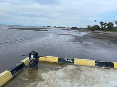 The image depicts a waterfront scene in Lancok, Indonesia, featuring a concrete pier with black and yellow markings. A black bollard stands on the pier, with a metal bar extending from it. The pier is wet and shows signs of wear and graffiti. Beyond the pier, a body of water stretches towards the horizon, with a sandy or muddy shoreline visible. The sky is overcast with clouds. Vegetation, including palm trees, is present in the distance along the shoreline. The overall scene is one of a functional, somewhat weathered, coastal area.