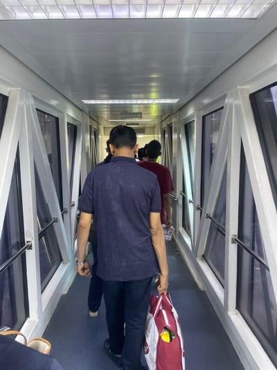 The image depicts a group of people walking through a jet bridge, likely at Sardar Vallabhbhai Patel International Airport in Ahmedabad, India. The jet bridge has a white metal frame and large glass panels on both sides, offering a view of the exterior which is not clearly visible due to reflections and the angle. The ceiling is illuminated by fluorescent lights arranged in a grid pattern.
In the foreground, a man wearing a dark blue short-sleeved shirt with a paisley pattern is walking away from the camera. He has a watch on his left wrist and is carrying a red and white reusable shopping bag in his right hand, which appears to contain some yellow items. Behind him, another man in a dark shirt is visible, and further ahead, a person in a red t-shirt is walking. The floor of the jet bridge is a dark blue carpet. The overall scene suggests passengers boarding or disembarking an aircraft.