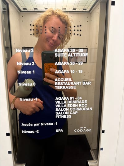 A blonde woman with her eyes closed and a smile on her face is taking a mirror selfie in what appears to be an elevator or a dressing room. She is wearing a blue tank top and black pants. She is holding a smartphone with a red pop socket and a green water bottle in her left arm. There are multiple lists of names and numbers on the mirror, including "Niveau 3", "AGAPA 30-39", "Niveau 2", "AGAPA 20-29", "Niveau 1", "AGAPA 10-19", "ACCUEIL", "RESTAURANT BAR", "TERRASSE", "Niveau -1", "AGAPA 01 - 04", "VILLA DESIRADE", "VILLA EDEN ROC", "SALON CORMORAN", "SALON CAP", "FITNESS", "Accès par Niveau -1", "Niveau -2", and "SPA BY CODAGE". The woman is wearing a watch on her left wrist. The background is the interior of the elevator or room, with a tiled ceiling and white walls.