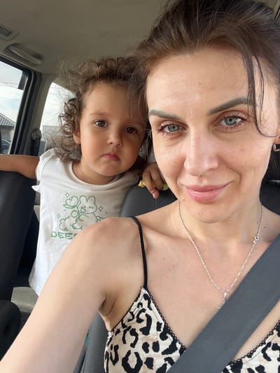 This is a selfie taken from inside a vehicle, possibly a car, with two subjects visible. A woman in the foreground is looking towards the camera, smiling, and wearing a necklace. She has light blue eyes and dark brown hair. To her left and slightly behind her, a young child is also visible, looking at the camera with a neutral expression. The child has curly brown hair and is wearing a white t-shirt with a green graphic print. They appear to be in the back seat of the car. The interior of the car is dimly lit, with a seatbelt visible across the woman's chest. The background shows a blurry view of the outdoors, including what appears to be a house with a dark roof and a cloudy sky. The image location is not specified in the provided information.