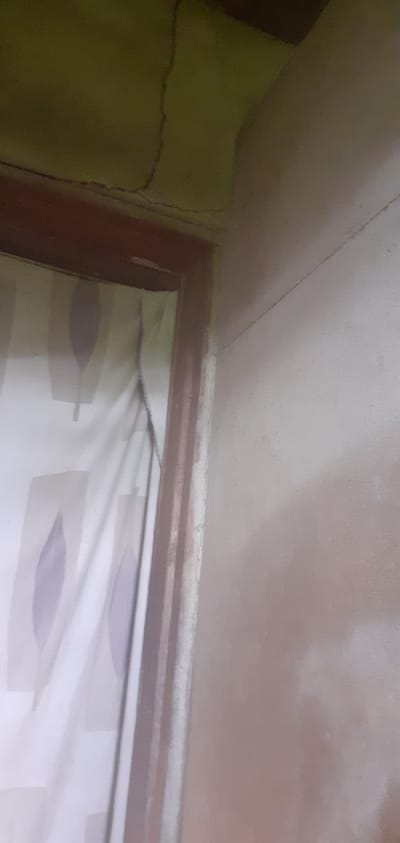 The image shows a corner of a room. The ceiling is a light beige color with a crack running across it. The wall on the left is light green with visible cracks. A dark brown wooden frame is visible at the corner, possibly of a window or a doorway. Inside the frame, a white curtain with light purple leaf-like patterns can be seen. The location is Alasia, Nigeria.