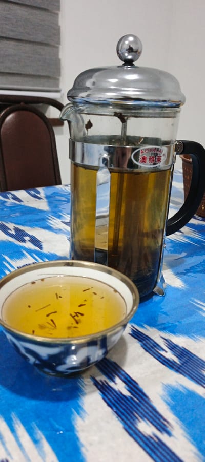 The image depicts an indoor scene in Moynaq, Uzbekistan, focusing on a prepared tea service on a patterned tablecloth. A clear glass French press with a chrome lid, frame, and black handle is positioned towards the center-right. It contains an amber-colored liquid with visible tea leaves compressed at the bottom. An oval white label with red Chinese characters, "奥雅莱" (AOYALAI), is affixed to the chrome band of the press. To the lower left, a small, white ceramic bowl with a gold rim and a blue and white patterned exterior holds a lighter amber liquid containing several small, dark, thread-like particulates floating on the surface. Both vessels rest on a table covered with a white tablecloth featuring a distinct blue Ikat-style pattern. The background is out of focus, revealing the upper portion of a brown upholstered chair and horizontal grey window blinds against a light-colored wall. No individuals, explicit actions, or interactions are visible.
