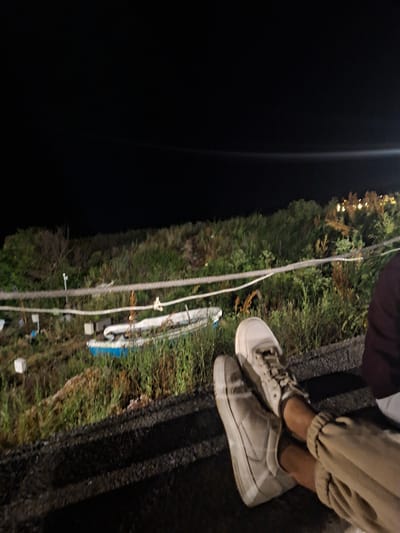 The image captures a nighttime scene in Tyulenovo, Bulgaria, focusing on a pair of legs and feet adorned with white sneakers and beige pants. The individuals are seated, with one leg resting atop the other. In the background, a small, overturned white and blue boat lies amidst dense green vegetation. A thick rope, likely a barrier, runs horizontally across the mid-ground, separating the foreground from the background. The sky is dark and cloudless, with distant lights hinting at a settlement beyond the natural landscape. The overall impression is one of peaceful contemplation in a coastal environment.