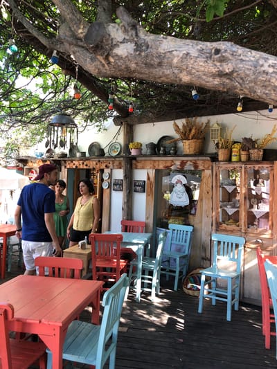 The image depicts an outdoor seating area of a cafe or restaurant in Kaş, Turkey, with a rustic and bohemian aesthetic. A large, gnarled tree with lush green foliage provides overhead shade, from which colorful string lights and a vintage-style hanging lamp are suspended. The seating area is furnished with a mix of brightly colored wooden tables and chairs, including coral-colored tables and chairs, and light blue chairs.

In the background, a charming wooden structure houses shelves filled with decorative items, kitchenware, and jars of dried goods. One section of the structure appears to be a display cabinet with glass doors, showcasing plates, glassware, and a pink teapot. A doorway leads into the interior of the establishment, visible through a glass panel that has a cutout sticker of a woman in traditional attire.

Three individuals are present in the scene. A man wearing a blue t-shirt and white shorts is standing on the left, facing two women. One woman, in a green dress, is looking towards him, while the other, in a yellow top and black shorts, is looking slightly away with an expressive gesture. The wooden floorboards of the outdoor seating area are visible throughout the foreground and mid-ground. The overall atmosphere is relaxed and inviting, suggesting a casual dining experience in a scenic location.