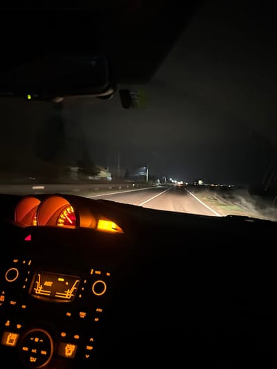 The image captures a nighttime view from the interior of a moving vehicle on a road in Okop, Bulgaria. The lower half of the frame prominently displays the vehicle's illuminated dashboard and center console. Two circular gauges, glowing orange, are visible in the instrument cluster, with one showing a needle indicating ongoing operation. The central console features an infotainment system displaying "AUX" and climate controls showing "23.0" degrees Celsius, all backlit in orange. A rearview mirror with a small green indicator light is positioned at the top center of the windshield.

Ahead, the two-lane road is illuminated by the vehicle's headlights and distant light sources, suggesting a continuation of the road or a nearby settlement. White lane markings are clearly visible on the dark asphalt. To the left of the road, a shoulder leads to indistinct structures, possibly buildings, in the distance, along with a utility pole. The right side of the road appears to be a verge with sparse, dimly lit vegetation. The sky is dark, consistent with nighttime conditions. No people are explicitly visible, but the vehicle's interior and moving perspective imply an occupant driving.