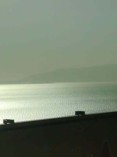 The image presents a serene, hazy landscape, capturing a wide body of water, likely Iznik Lake in Turkey, under a diffused sky. The perspective appears to be slightly elevated, looking out over the water towards distant landmasses.

In the immediate foreground, a dark, solid architectural structure, resembling a wall or balcony railing, extends across the bottom of the frame. Two small, dark, rectangular objects are discernible atop this structure, hinting at either functional elements or items placed upon it.

Beyond this foreground element, the expansive water surface dominates the mid-ground. It is intensely illuminated by direct sun reflections, creating a dazzling, shimmering texture that covers the entire visible area of the lake. This strong glint suggests a bright day, possibly midday or late afternoon.

Further in the distance, a faint, light-colored line delineates a distant shoreline or landmass. Behind this, the soft, undulating forms of mountains or hills rise gently, their contours softened and muted by atmospheric haze. The sky above is uniformly hazy, appearing as a pale, greenish-grey expanse that blends seamlessly with the distant land, creating a sense of depth and tranquility.

No people or specific activities are visible, emphasizing the natural grandeur and vastness of the scene. The overall impression is one of calm, ethereal beauty, with the hazy light giving the landscape a dreamy quality. There is no visible text in the image.