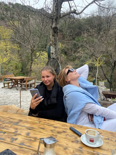 Two young women are seated at a wooden table in an outdoor setting. One woman, wearing a black jacket, is holding a smartphone and taking a selfie, while the other woman, dressed in a white shirt and a light blue hoodie, is leaning back with her head resting on her hands. On the table is a cup of coffee, a lighter, and a glass container. In the background, there are trees, and a partial view of a mountain. The location appears to be a cafe or outdoor seating area, and the scene is set in the vicinity of Krapina, Montenegro, considering the surrounding vegetation and the architectural style of the outdoor furniture and landscape.