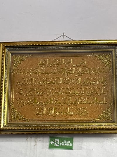 The image shows a framed Islamic calligraphy hanging on a white wall. The calligraphy is in Arabic script and appears to be embossed on a textured, golden background. The frame is ornate and golden. Below the framed artwork, there is a green and white "JALUR EVAKUASI" (evacuation route) sign with an arrow pointing to the left. The environment appears to be indoors, possibly a public building or a place of worship. There are no people or animals visible in the image. The lighting is moderate, casting some shadows on the wall. The city of Julicotmeurak, Indonesia, is mentioned in the prompt, but there are no visual cues in the image to confirm this specific location.